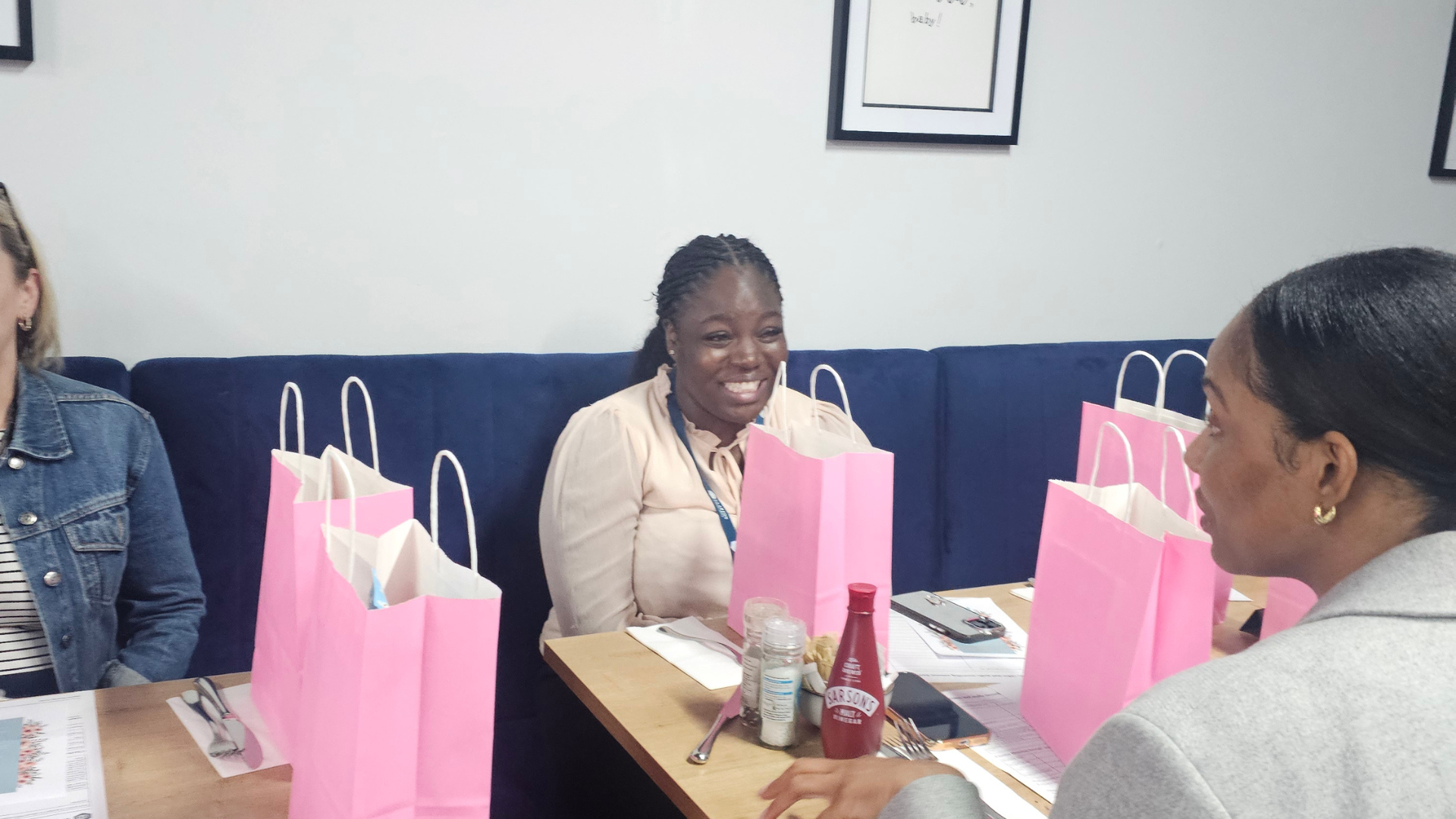 People sitting opposite each other at a table with pink gift bags, printed materials and drinks at a community brunch.
