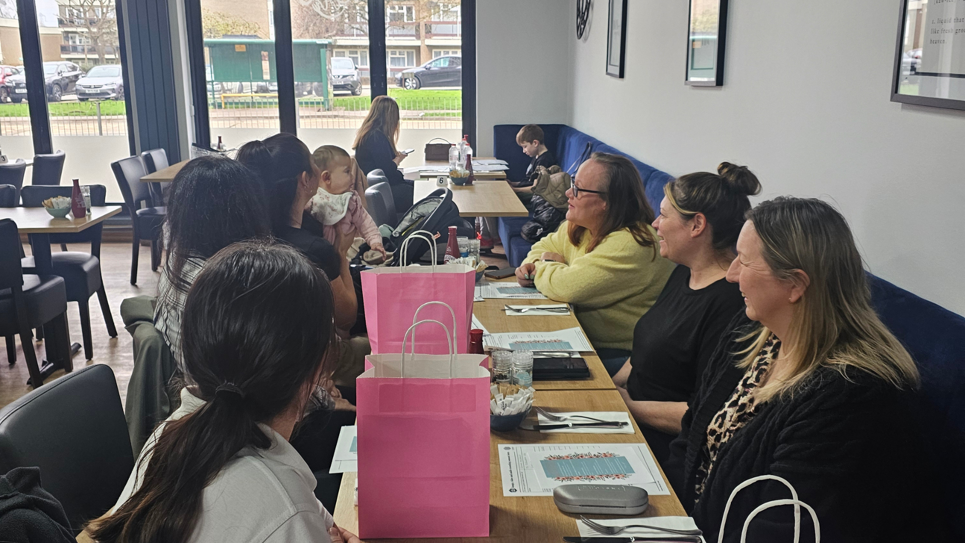 A group of people seated at tables during a community brunch, with printed information sheets and gift bags visible.