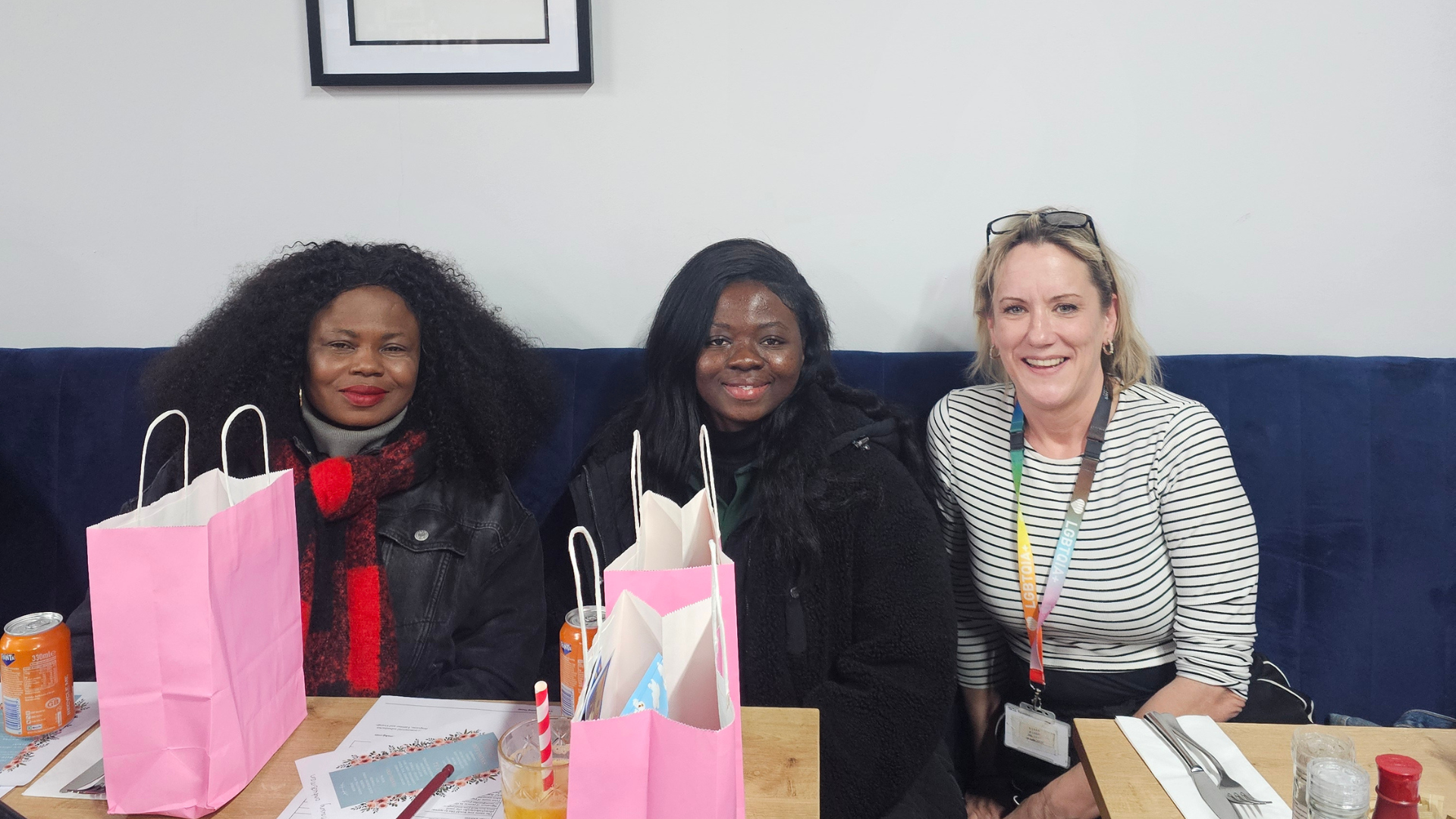 Three people seated at a table with pink gift bags, printed leaflets and drinks at a community brunch.