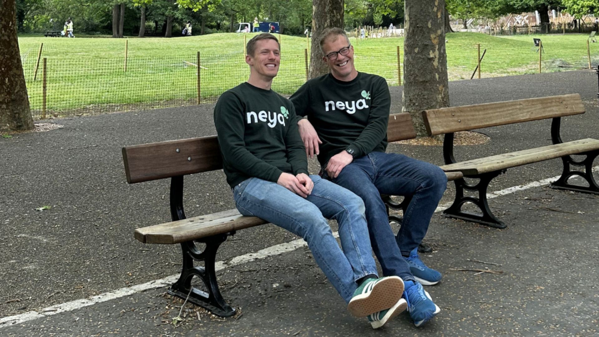 Two people wearing matching “neya” sweatshirts sit on a wooden park bench beside a paved path, with trees, fencing, and open green space in the background.