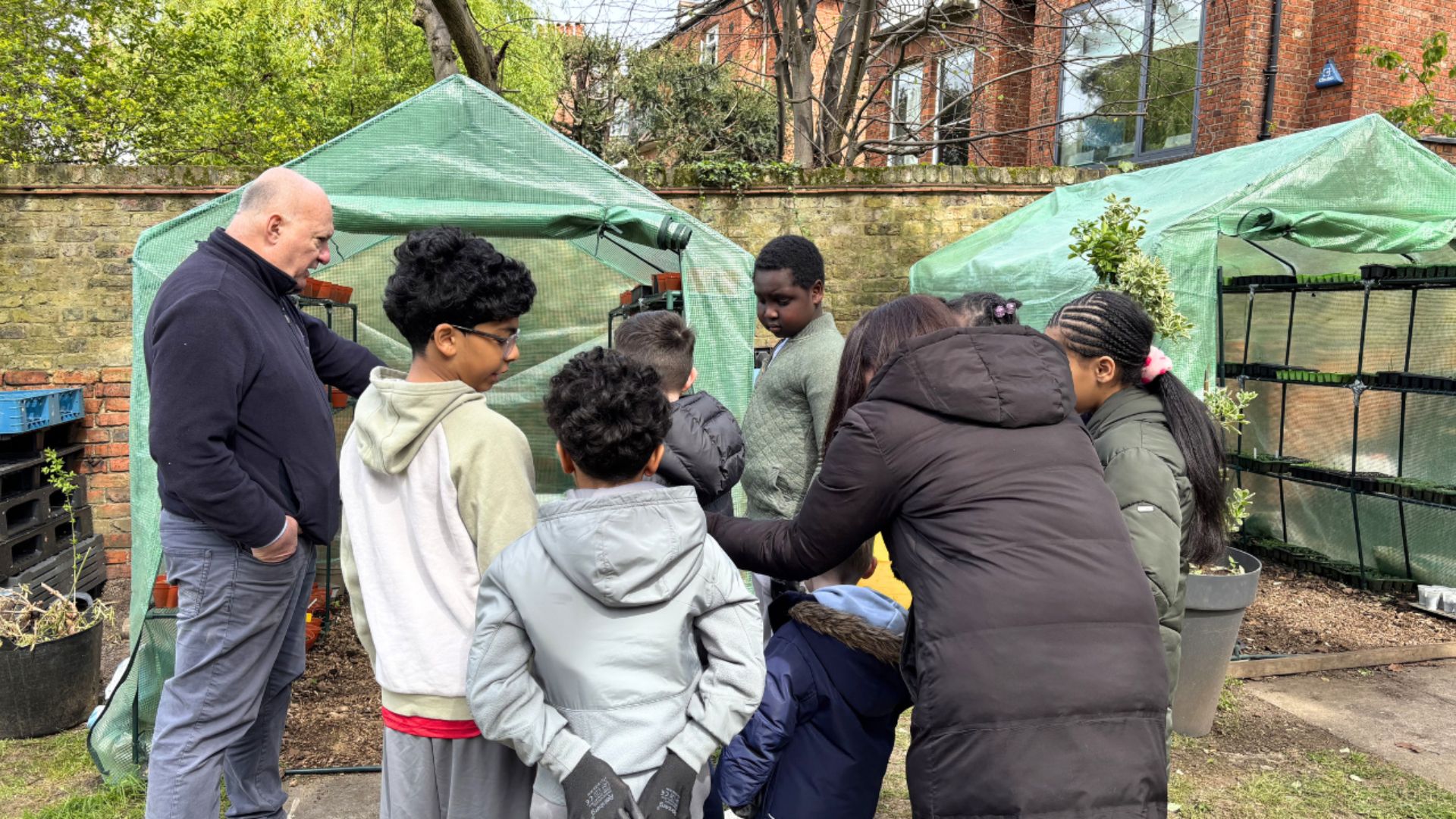 Children and adults gathered around raised beds and small greenhouses during a community gardening activity.