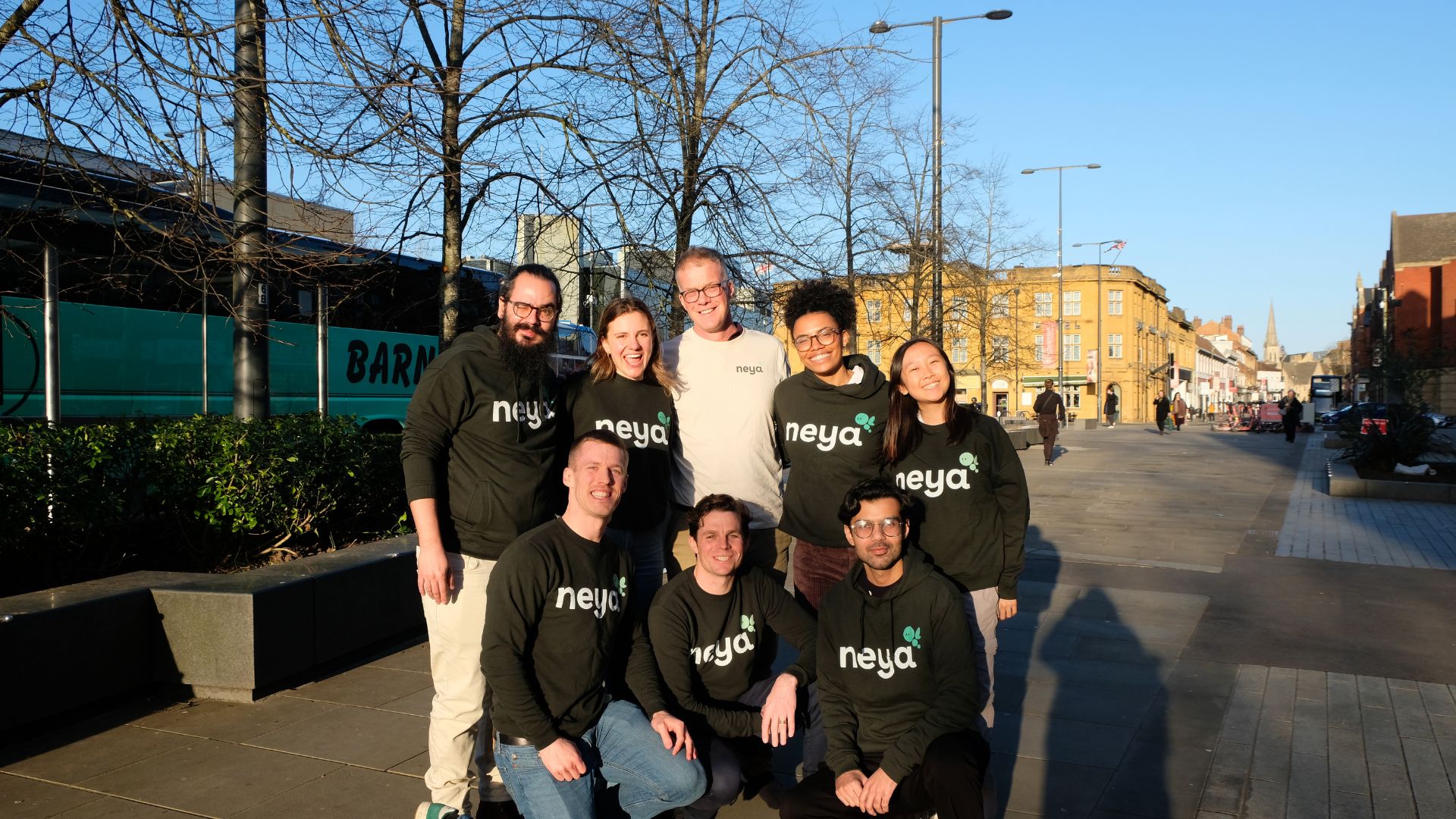 A group of nine people wearing “neya” branded tops pose together on a sunlit town street, with buildings, trees, and pedestrians behind them.