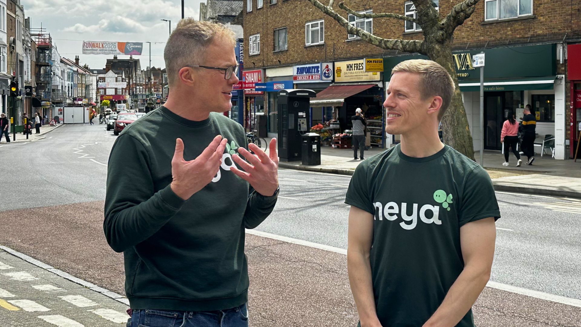 Two people wearing “neya” t-shirts stand on a high street and talk, with shops, parked cars, and a large tree lining the road in the background.