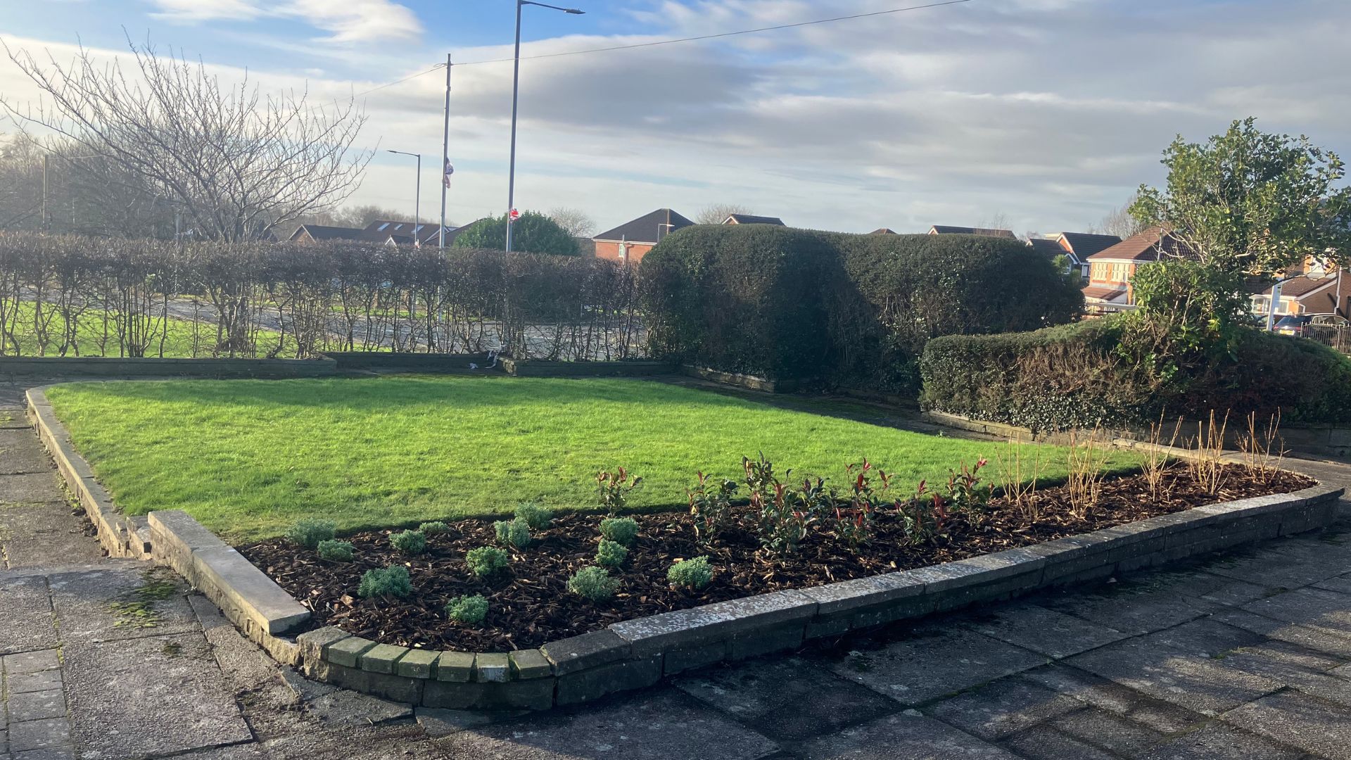 Small landscaped green space in a residential area with a central grass lawn, low brick-edged planting bed with shrubs in the foreground, neatly trimmed hedges along the perimeter