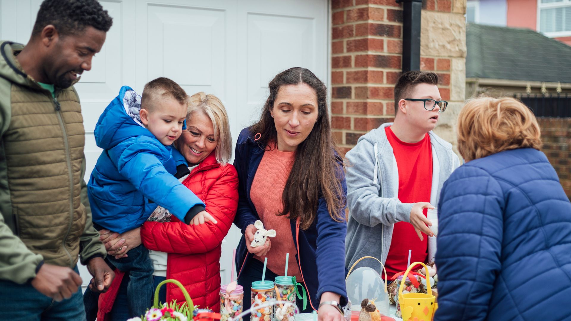 People gathered around an outdoor table with colorful baskets, jars, and decorations in front of a brick house.